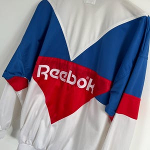 Op de afbeelding: Een vintage Reebok trainingsjack in wit, blauw en rood. De jas heeft een colorblock-ontwerp met het witte Reebok-logo op de borst. Het heeft een hoge kraag en een ritssluiting over de hele lengte.