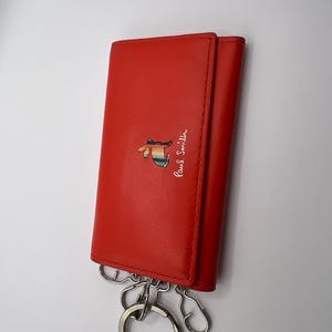 Puede incluir: Un estuche para llaves de cuero rojo con un llavero y ganchos plateados. El estuche tiene un gráfico colorido y el logotipo de Paul Smith en escritura blanca. El estuche está cerrado y tiene un cierre a presión.