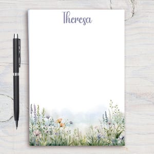 May include: A notepad with the name "Theresa" in script at the top. The bottom of the notepad features a watercolor illustration of wildflowers in shades of purple, orange, and green. A black pen rests on the left side.