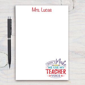 Op de afbeelding: Wit notitieblok met rode tekst die bovenaan "Mrs. Lucas" zegt. De onderkant van het notitieblok heeft een kleurrijke afbeelding die "Don't Make Me Use My Teacher Voice" zegt.