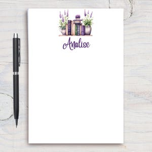 May include: A white notepad with a purple floral design and the name Analise printed on it. The design features a stack of books with lavender plants on either side.