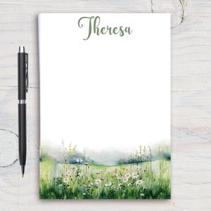 Op de afbeelding: Notitieblok met de naam "Theresa" in groene letters bovenaan. De onderkant van de pagina toont een aquarel van een veld met madeliefjes en wilde bloemen. Een zwarte pen ligt aan de linkerkant.