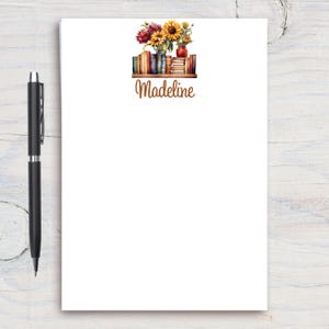 May include: Personalized notepad with a floral design featuring sunflowers and other flowers in a vase on a shelf with books. The name Madeline is printed in brown text below the image.