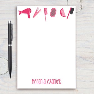 May include: White notepad with a pink border and the text "Megan Alexander" at the bottom. The top border features pink hair styling tools, including a hairdryer, curling iron, comb, brush, and a hair pick.