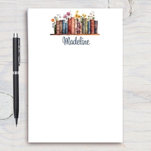 Pode incluir: Um bloco de notas branco com um design floral e uma fileira de livros coloridos em uma prateleira. O nome "Madeline" está escrito em azul cursivo na parte inferior do bloco de notas.