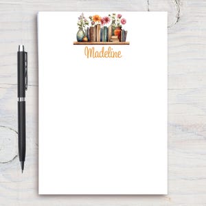May include: A white notepad with a floral design and the name "Madeline" printed on it. The design features a shelf with books, vases, and flowers.