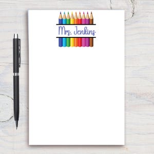 May include: White notepad with a black pen on a wooden surface. The notepad features a colorful pencil design and the name "Mrs. Jenkins" in blue script. The pencils are arranged in a rainbow pattern.
