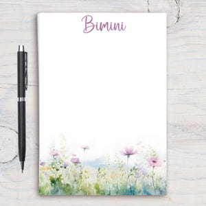 Może przedstawiać: Biały notatnik z napisem "Bimini" w fioletowym skrypcie na górze. Dół notatnika zdobi akwarelowy wzór kwiatowy z różowymi i fioletowymi kwiatami. Czarny długopis spoczywa po lewej stronie notatnika.