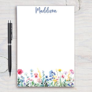 May include: White notepad with the name "Maddison" in blue script at the top. The bottom edge features a watercolor floral design with pink, blue, yellow, and purple flowers. A black pen rests on the left side.