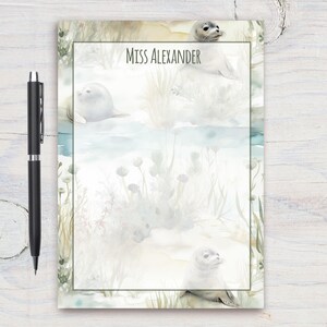 Può includere: Un blocco note con un design ad acquerello di foche e alghe marine. Il blocco note ha uno sfondo blu chiaro con un bordo bianco. Il testo "Miss Alexander" è stampato nella parte superiore del blocco note.