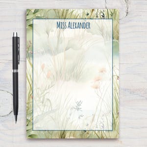 Può includere: Un blocco note con un design ad acquerello di erba verde e fiori. Il blocco note ha un bordo blu e il testo "Miss Alexander" in alto.