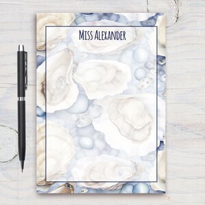 Può includere: Un blocco note con un design ad acquerello di conchiglie di ostriche in tonalità di bianco e blu. Il testo "Miss Alexander" è stampato sulla parte anteriore del blocco note.