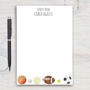 Op de afbeelding: Een wit notitieboekje met de tekst "A Note From Coach Agassi" bovenaan. De onderkant van het notitieboekje heeft een rij kleurrijke sportballen, waaronder een basketbal, een honkbal, een volleybal, een voetbal en een tennisbal.