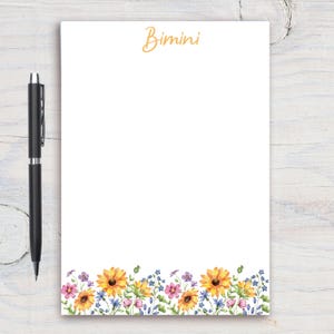 May include: White notepad with a floral border at the bottom. The border features yellow sunflowers, pink and purple flowers, and green leaves. The top of the notepad has the word "Bimini" written in orange.