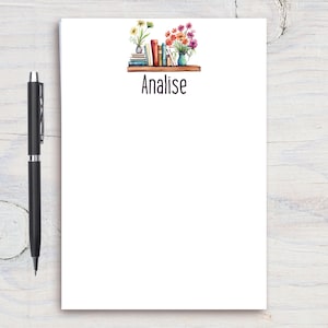 May include: A white notepad with a floral and book illustration and the name Analise printed on it.