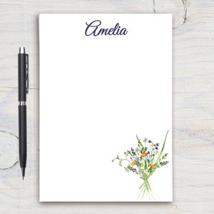 Pode incluir: Bloco de notas branco com o nome Amelia impresso em itálico roxo no topo. O bloco de notas tem um design floral na parte inferior com um buquê de flores silvestres brancas, roxas e amarelas.