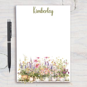 May include: A personalized notepad with the name "Kimberley" in green script at the top. The bottom of the notepad features a watercolor illustration of various potted flowers in shades of pink, purple, yellow, and white. A black pen rests on the left.