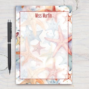 Può includere: Un blocco note bianco con un bordo rosso e un disegno ad acquerello di stelle marine in tonalità di rosa, blu e arancione. Il testo "Miss Murfin" è stampato in rosso nella parte superiore del blocco note.
