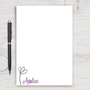 Pode incluir: Um bloco de notas branco com um design simples de flor preta e o nome Analise escrito em letra cursiva roxa.