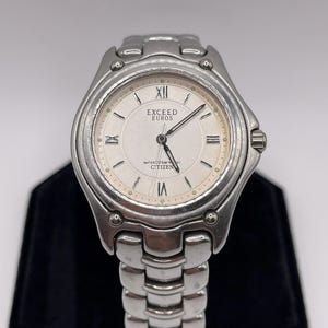 May include: A silver-toned Citizen Exceed Euros wristwatch. The watch features a cream-colored face with Roman numeral hour markers and the text "EXCEED EUROS". The watch band is made of linked metal. The watch is water resistant to 20 bar.