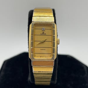 May include: A gold-toned Lorus Quartz watch with a rectangular face and a gold-colored band. The watch face has a small diamond accent and the word "QUARTZ" is printed on the face. The watch is displayed on a black surface.