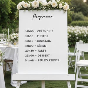Può includere: Cavalletto bianco con un cartello con la scritta "Programme" e un programma di nozze. Il cartello è decorato con fiori bianchi. Il programma include gli orari per la cerimonia, le foto, il cocktail, la cena, la festa, il dessert e i fuochi d'artificio.