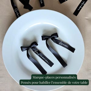 May include: Black satin ribbon place cards with white script names and bow details. The place cards are arranged on a white plate. The text on the image reads "Marque-places personnalis&eacute;s Pens&eacute;s pour habiller l'ensemble de votre table."