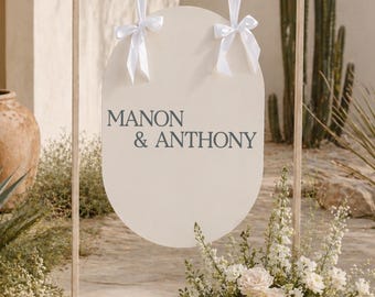 May include: A cream-colored wedding sign with the names "MANON & ANTHONY" in dark gray lettering. The sign is oval-shaped and hangs from a wooden frame with white ribbon bows. The scene includes flowers and desert plants.