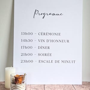 Peut inclure: Un panneau de programme de mariage rectangulaire blanc avec du texte noir indiquant l'horaire : 13h00 CÉRÉMONIE, 14h30 VIN D'HONNEUR, 17h00 DÎNER, 21h00 SOIRÉE et 23h00 ESCALE DE MINUIT. Une bougie pilier blanche et un vase en verre avec du sable et une fleur séchée sont posés à côté.