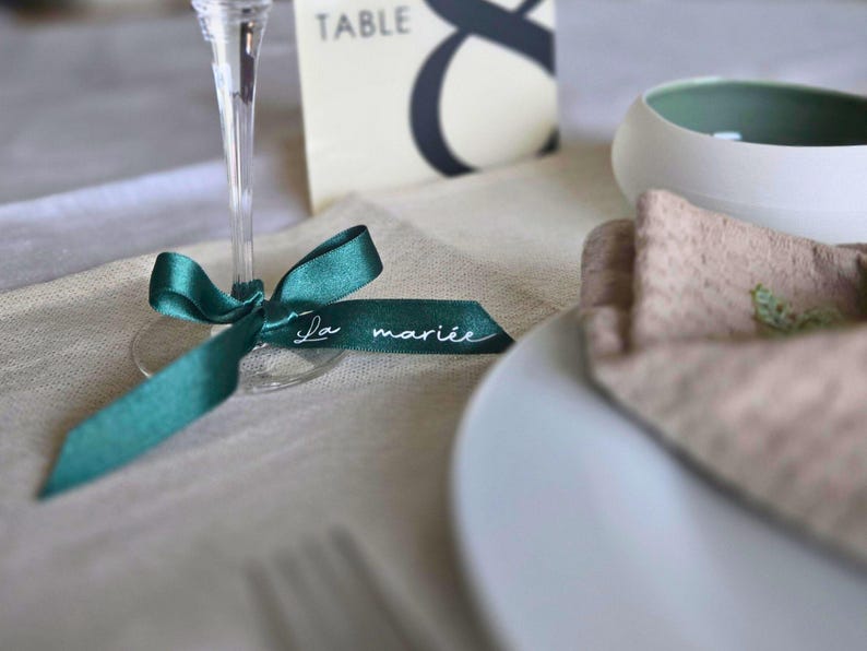 May include: A teal ribbon tied around a champagne glass, with the words "La mari&eacute;e" in white script. A white plate, a folded napkin, and a table number card are also visible, suggesting a wedding or event setting.