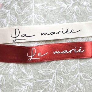 May include: Two ribbons with French text. One is cream-colored with "La mari&eacute;e" in black script. The other is red with "Le mari&eacute;" in white script. Both ribbons have angled ends and are set against a floral background.