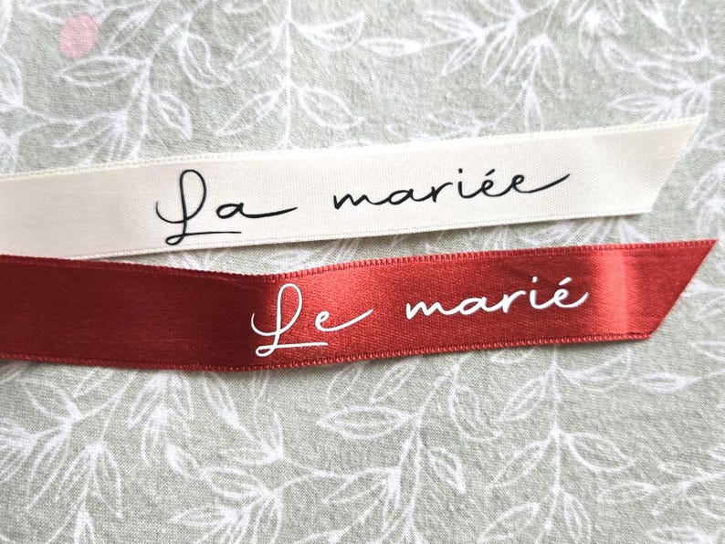 May include: Two ribbons with French text. One is cream-colored with "La mari&eacute;e" in black script. The other is red with "Le mari&eacute;" in white script. Both ribbons have angled ends and are set against a floral background.