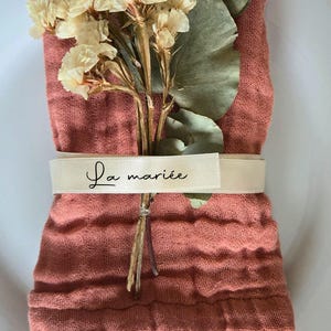 May include: A folded coral-colored linen napkin is adorned with dried flowers and greenery. A cream-colored ribbon encircles the napkin, displaying the text "La mari&eacute;e" in elegant script. The arrangement sits on a white plate.