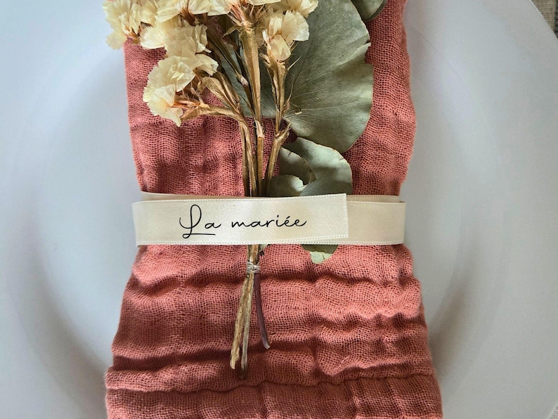 May include: A folded coral-colored linen napkin is adorned with dried flowers and greenery. A cream-colored ribbon encircles the napkin, displaying the text "La mari&eacute;e" in elegant script. The arrangement sits on a white plate.