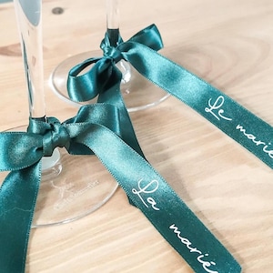 May include: Two champagne flutes with teal satin ribbons tied in bows. Each ribbon is printed with the words "Le mari&eacute;" in white script. The flutes are on a light wood surface.