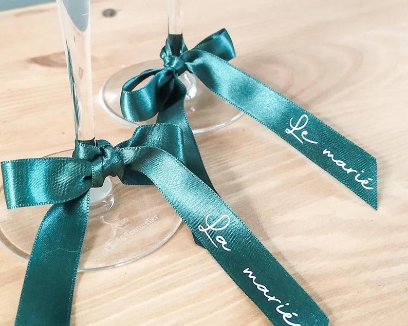 May include: Two champagne flutes with teal satin ribbons tied in bows. Each ribbon is printed with the words "Le mari&eacute;" in white script. The flutes are on a light wood surface.