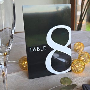 Peut inclure: Un carton de numéro de table noir avec un grand chiffre huit blanc. Le mot « TABLE » est imprimé en blanc. Des guirlandes lumineuses décoratives avec des sphères dorées sont en arrière-plan.