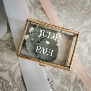 May include: A rectangular glass box with gold trim, containing a gray stone and white flowers. The box has the names "JULIE" and "PAUL" with a heart symbol in between. The box is on a white ribbon and a pink ribbon.