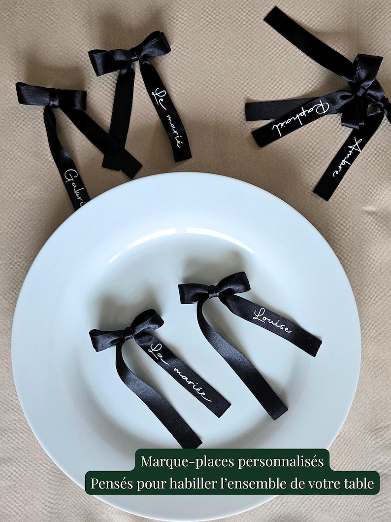 May include: Black satin ribbon place cards with white script names and bow details. The place cards are arranged on a white plate. The text on the image reads "Marque-places personnalis&eacute;s Pens&eacute;s pour habiller l'ensemble de votre table."