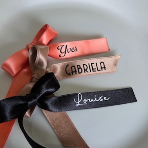 May include: Close-up of three satin ribbons with names printed on them. The ribbons are in coral, tan, and black. The names are "Yves", "Gabriela", and "Louise". The ribbons are tied into bows. The text "Couleurs et &eacute;critures au choix" is at the bottom.
