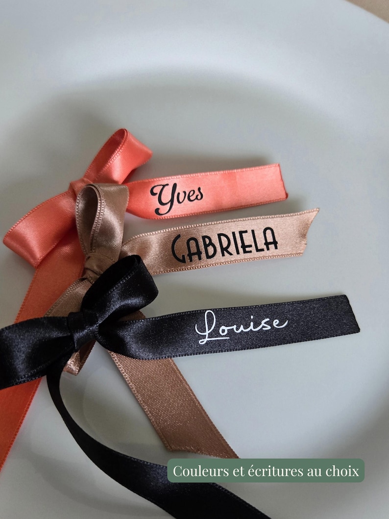 May include: Close-up of three satin ribbons with names printed on them. The ribbons are in coral, tan, and black. The names are "Yves", "Gabriela", and "Louise". The ribbons are tied into bows. The text "Couleurs et &eacute;critures au choix" is at the bottom.