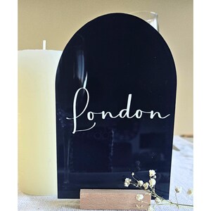 May include: A black acrylic sign with an arched top, featuring the word "London" in white script. The sign is held by a small wooden base. A white candle and small white flowers are in the background.