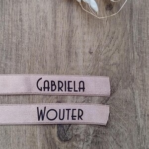 May include: Two light pink ribbons with black text. The top ribbon reads "GABRIELA" and the bottom ribbon reads "WOUTER". The ribbons are on a wooden surface.