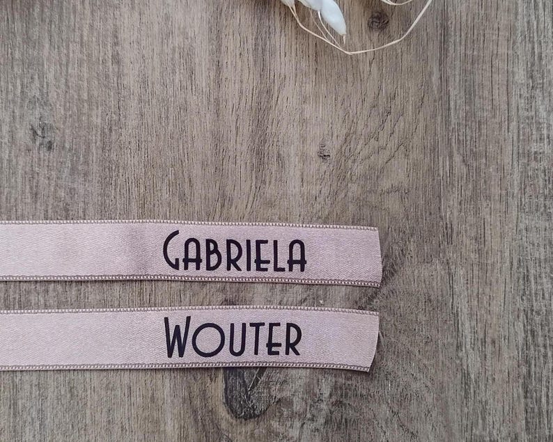 May include: Two light pink ribbons with black text. The top ribbon reads "GABRIELA" and the bottom ribbon reads "WOUTER". The ribbons are on a wooden surface.