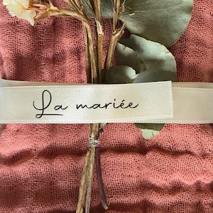 May include: A bundle of dried flowers and greenery wrapped in a coral-colored fabric. A cream-colored ribbon encircles the bundle, with the words "La mari&eacute;e" printed in black script. The arrangement suggests a wedding or celebratory theme.