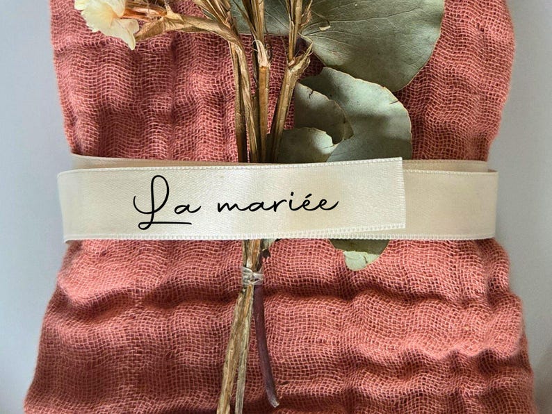 May include: A bundle of dried flowers and greenery wrapped in a coral-colored fabric. A cream-colored ribbon encircles the bundle, with the words "La mari&eacute;e" printed in black script. The arrangement suggests a wedding or celebratory theme.