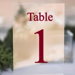 Peut inclure: Panneau de table en acrylique transparent avec le texte bordeaux. Le mot "Table" est au-dessus du chiffre "1". Le panneau est posé sur une base blanche. L'arrière-plan est flou.