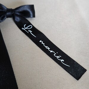 May include: A black satin ribbon bow with the text "La mari&eacute;e" in white script. The ribbon is tied in a bow and is set against a neutral background.
