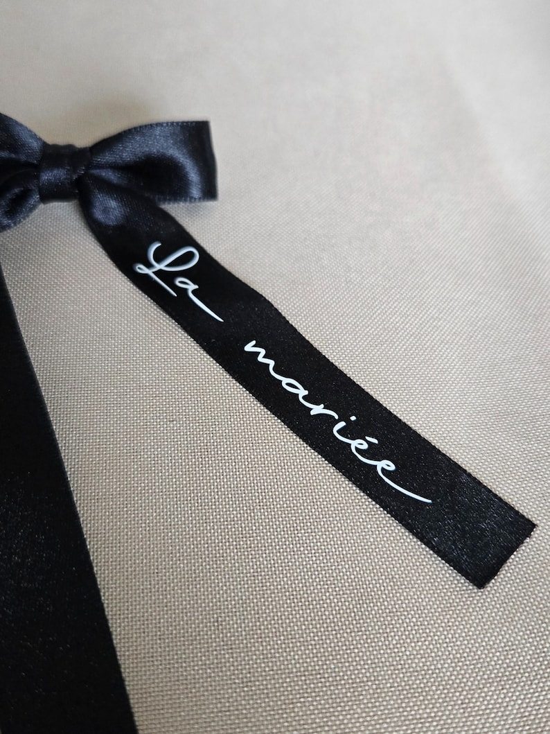 May include: A black satin ribbon bow with the text "La mari&eacute;e" in white script. The ribbon is tied in a bow and is set against a neutral background.