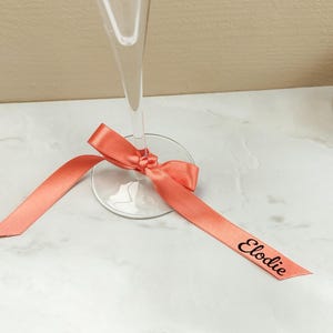 May include: A champagne flute with a coral ribbon tied around the base. The ribbon has the name "Elodie" printed on it in black. The glass is on a white marble surface.